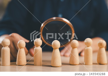 Magnifying glass examines wooden people in a row, representing recruitment, candidate search, employee screening, talent discovery, hiring, assessment, evaluation, and human resources management. 143543288