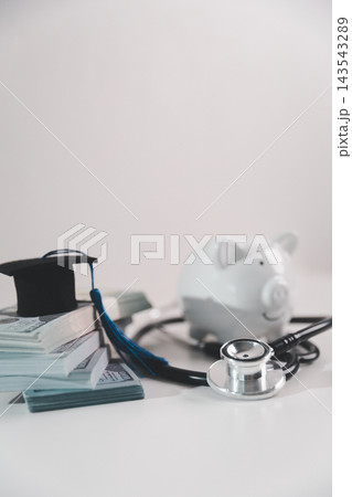 Graduation cap on stacks of money with stethoscope and piggy bank, representing medical education funding, tuition savings, student finance and investment in a future healthcare career. 143543289