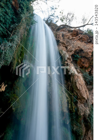 Tall Silky Waterfall Down a Mossy Rock Face. A tall silky waterfall pours down a mossy rock face amid green trees in the Monasterio de Piedra natural park in Aragon, Spain, a serene vertical long expo Tall Silky Waterfall Down a Mossy Rock Face. A tall silky waterfall pours down a mossy rock face amid green trees in the Monasterio de Piedra natural park in Aragon, Spain, a serene vertical long expo 143543820