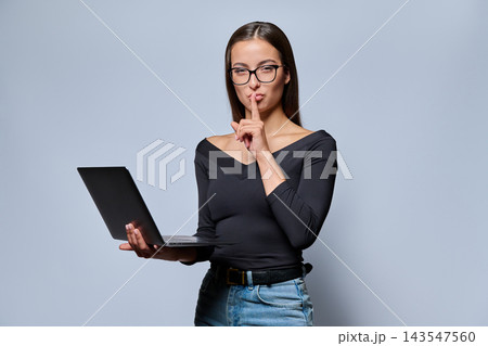 Young woman in glasses holding laptop and showing silence sign in studio Young woman in glasses holding laptop and showing silence sign in studio 143547560