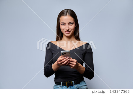 Smiling young woman holding smartphone and looking at camera in studio Smiling young woman holding smartphone and looking at camera in studio 143547563