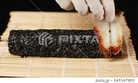 Chef adding grilled unagi eel to black rice sushi roll on bamboo mat, preparing Japanese seafood cuisine Chef adding grilled unagi eel to black rice sushi roll on bamboo mat, preparing Japanese seafood cuisine 143547903