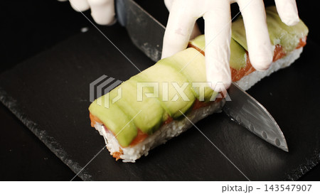 Chef's gloved hands slicing a sushi roll featuring fresh avocado, salmon, and sticky rice on a dark slate board Chef's gloved hands slicing a sushi roll featuring fresh avocado, salmon, and sticky rice on a dark slate board 143547907