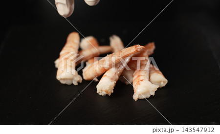 Chef's gloved hand dropping cooked shrimp onto a dark slate plate, preparing a seafood dish Chef's gloved hand dropping cooked shrimp onto a dark slate plate, preparing a seafood dish 143547912