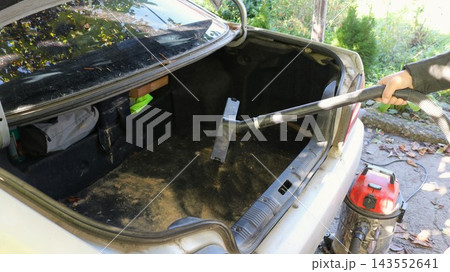The process of cleaning the open trunk of a white passenger car. A man's hand holds the hose of a powerful industrial vacuum cleaner, removing heavy dirt and wood shavings from the mat. 143552641