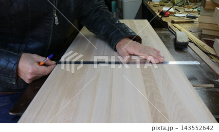 The hands of a craftsman in a leather jacket are marking out a wooden workpiece. A long steel ruler lies on the smooth, light-colored panel, and a mark is being made along it with a pen. 143552642