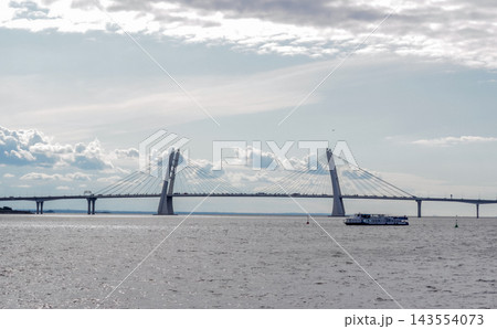 Modern cable-stayed bridge over wide river with passenger boat under cloudy sky Modern cable-stayed bridge over wide river with passenger boat under cloudy sky 143554073