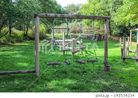 A woodland playground with rustic wooden climbing frames, ropes, and a nest swing sits on a lush green lawn, surrounded by dense trees and a picnic area under a leafy canopy in the soft daylight. 143554254