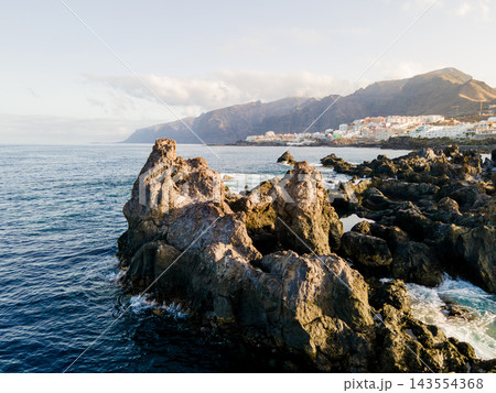Jagged lava spires rise from the sea below distant cliffs. Sharp volcanic spires break the surface in the foreground. Behind them the town of Los Gigantes and its vast basalt cliffs recede into soft m 143554368