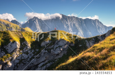 Two hikers walking along a grassy mountain ridge, rocky peaks Two hikers walking along a grassy mountain ridge, rocky peaks 143554853
