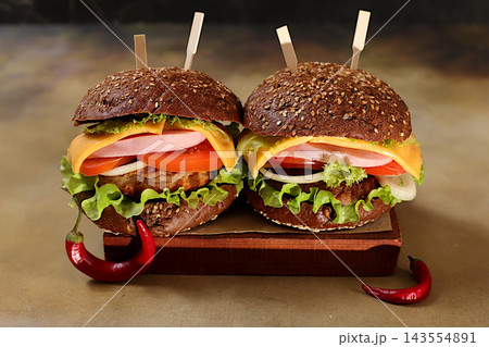 Two double cheeseburgers on whole-grain sesame seed buns featuring fresh lettuce, tomato, cream cheese, chili pepper, bacon, onion and a patty served on a wooden tray, food delivery concept, menu Two double cheeseburgers on whole-grain sesame seed buns featuring fresh lettuce, tomato, cream cheese, chili pepper, bacon, onion and a patty served on a wooden tray, food delivery concept, menu 143554891