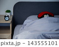 There is a black alarm clock on a wooden bedside table, and in the background a brown bear in a red mask sleeps wrapped in a white blanket under a gray headboard. Concept: the toy aesthetics of sleep 143555101