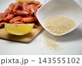 On a white background, there is a bowl of rice, a wooden board with frozen shrimp, a slice of lemon, and a few grains of cereal spilled onto the countertop. High quality photo 143555102