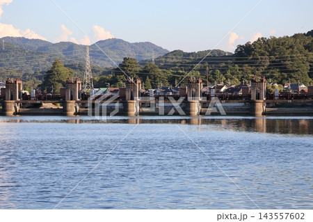 青い水面の川と洗堰の水門と緑の樹々の森と住宅街と山並みの雲と青空の風景 青い水面の川と洗堰の水門と緑の樹々の森と住宅街と山並みの雲と青空の風景 143557602