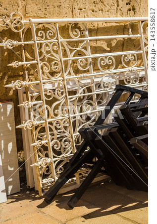 A vertical shot showing intricate vintage white metal scrolls and pointed fence spikes stacked alongside folded black furniture frames on stone paving. High quality photo A vertical shot showing intricate vintage white metal scrolls and pointed fence spikes stacked alongside folded black furniture frames on stone paving. High quality photo 143558617