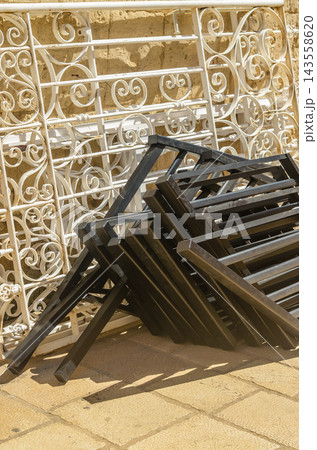 A close-up shot of folded black metal furniture frames stacked neatly in front of ornate, vintage white wrought iron balcony railings on a stone pavement.. High quality photo A close-up shot of folded black metal furniture frames stacked neatly in front of ornate, vintage white wrought iron balcony railings on a stone pavement.. High quality photo 143558620