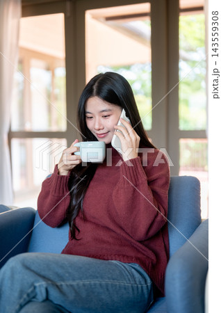 Young Asian woman holding and sipping coffee during smartphone call on sofa in a living room or cafe Young Asian woman holding and sipping coffee during smartphone call on sofa in a living room or cafe 143559308