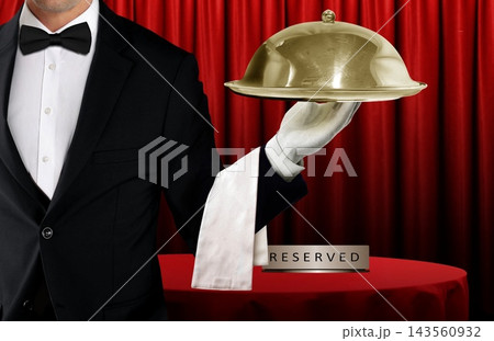 Waiter in black tuxedo holding covered food platter with a cloche in front of reserved table and red curtain Waiter in black tuxedo holding covered food platter with a cloche in front of reserved table and red curtain 143560932
