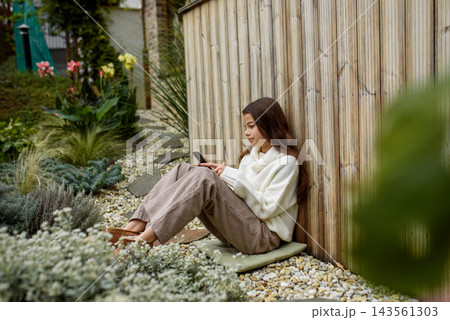 Young girl sitting on ground outdoors, holding smartphone and scrolling. 143561303