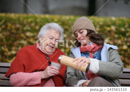 Portrait of a granddaughter and grandmother sitting on bench in autumn park, drinking coffee and laughing together. Portrait of a granddaughter and grandmother sitting on bench in autumn park, drinking coffee and laughing together. 143561310