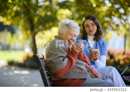Elderly woman enjoying cup of coffee with her nurse in the park during warm autumn day. Elderly woman enjoying cup of coffee with her nurse in the park during warm autumn day. 143561322