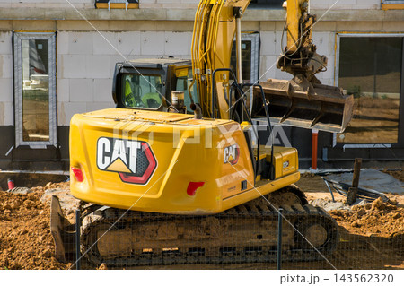 An excavator is working on a construction site, moving soil and debris during the day. The heavy machinery is positioned next to an incomplete building structure with exposed walls. 143562320