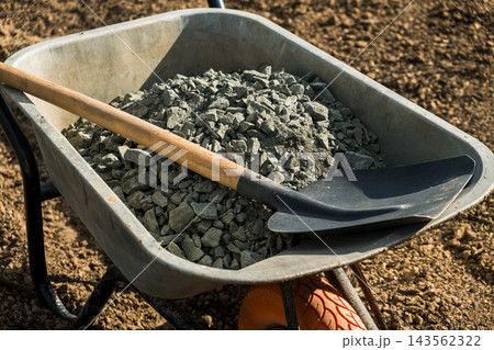 Crushed gravel fills a wheelbarrow beside a shovel on bare soil at an outdoor construction site under bright sunlight, closeup. Crushed gravel fills a wheelbarrow beside a shovel on bare soil at an outdoor construction site under bright sunlight, closeup. 143562322