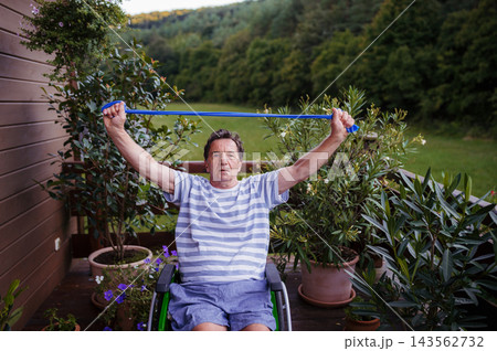 Senior man in wheelchair exercising with resistance band on terrace. Senior man in wheelchair exercising with resistance band on terrace. 143562732