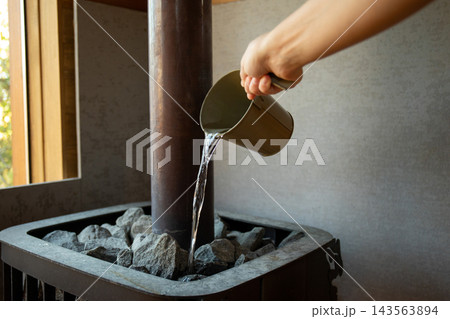 Person adds water to hot stones in sauna. Relaxation and wellness concept Person adds water to hot stones in sauna. Relaxation and wellness concept 143563894