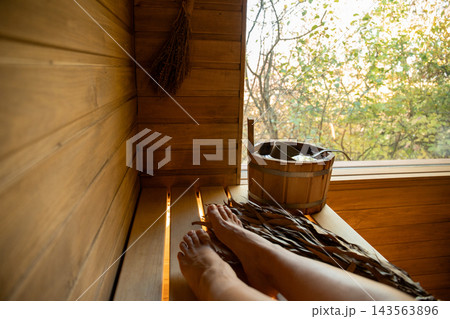 Feet resting on wooden bench inside sauna while enjoying serene nature view Feet resting on wooden bench inside sauna while enjoying serene nature view 143563896