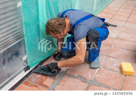 Worker grouting outdoor terracotta tiles beside covered glass storefront Worker grouting outdoor terracotta tiles beside covered glass storefront 143565056
