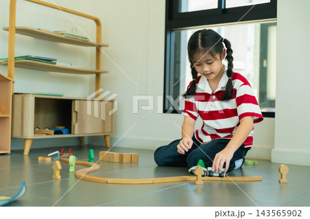 Young girl playing with wooden toy train set on floor in bright modern living room with shelves Young girl playing with wooden toy train set on floor in bright modern living room with shelves 143565902