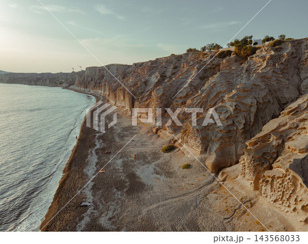 Aerial shot flying sideways along the rocky cliffs of Eros Beach, Santorini. Aerial view gliding alongside the rocky cliffs and shimmering blue waters of the Mediterranean Sea at a Santorini beach, Gr 143568033