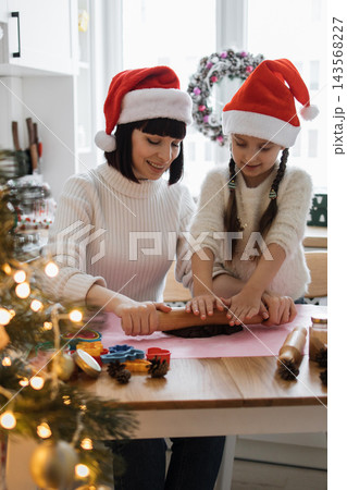 A mother and daughter wearing Santa hats are happily rolling out cookie dough together in a festive kitchen setting A mother and daughter wearing Santa hats are happily rolling out cookie dough together in a festive kitchen setting 143568227