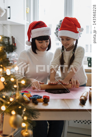 A mother and daughter wearing Santa hats are making Christmas cookies together in a cozy kitchen setting A mother and daughter wearing Santa hats are making Christmas cookies together in a cozy kitchen setting 143568228