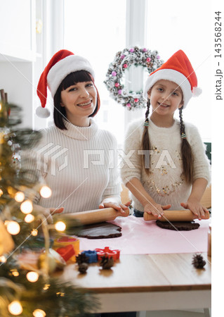A mother and daughter wearing Santa hats happily bake Christmas cookies together in a festive kitchen setting 143568244