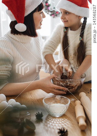 A mother and daughter wearing Santa hats joyfully bake Christmas cookies together, mixing dough in a bowl 143568287
