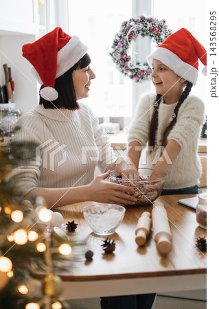A mother and daughter wearing Santa hats joyfully bake Christmas cookies together in a cozy kitchen setting 143568295