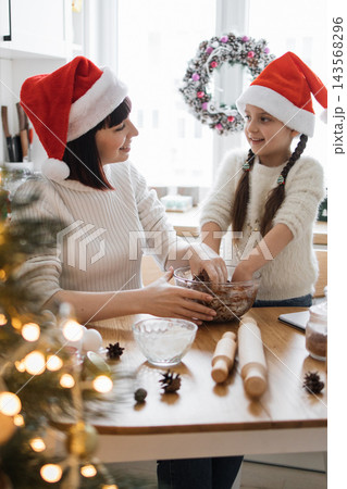 A mother and daughter wearing Santa hats joyfully bake Christmas cookies together in a festive kitchen 143568296