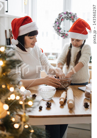 A Caucasian mother and daughter wearing Santa hats joyfully bake Christmas cookies together in a festive kitchen setting A Caucasian mother and daughter wearing Santa hats joyfully bake Christmas cookies together in a festive kitchen setting 143568297