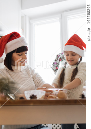 A mother and daughter wearing Santa hats are happily baking Christmas cookies together in a bright kitchen 143568298