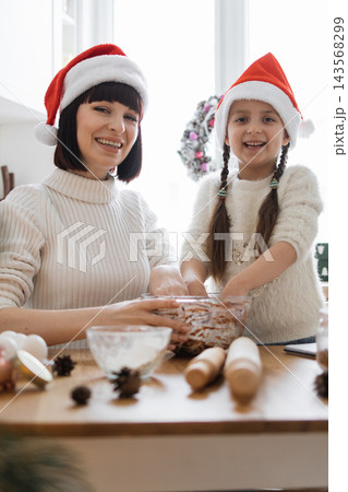 A mother and daughter wearing Santa hats are happily baking Christmas treats together in a cozy kitchen setting 143568299