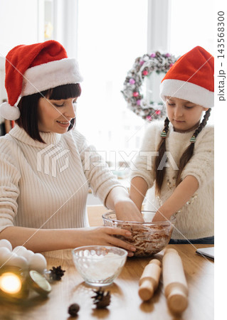 A mother and daughter wearing Santa hats joyfully bake Christmas cookies together in a cozy kitchen setting A mother and daughter wearing Santa hats joyfully bake Christmas cookies together in a cozy kitchen setting 143568300