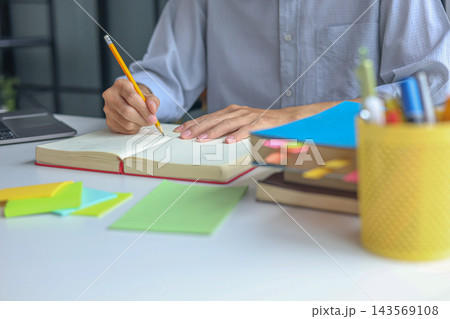 University Student Studying and Writhing in Notebook Preparing for Exam with Textbook on the Table at Home. 143569108