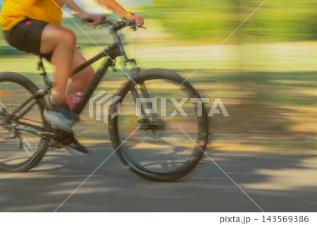 Cyclist on Mountain Bike, Motion Blur. Cyclist wearing yellow shirt rides mountain bike along sunlit paved path with motion blur. Dynamic cycling scene, movement, outdoor recreation Cyclist on Mountain Bike, Motion Blur. Cyclist wearing yellow shirt rides mountain bike along sunlit paved path with motion blur. Dynamic cycling scene, movement, outdoor recreation 143569386