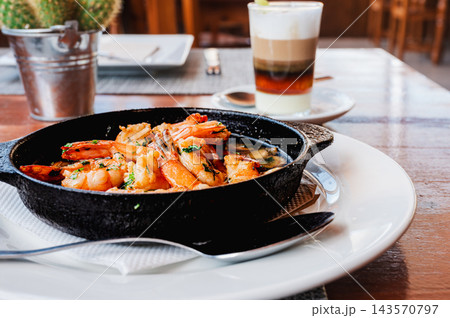 Garlic prawns and coffee on a polished restaurant table. A dish of garlic prawns and a barraquito coffee sit on a polished wooden table in a Canarian restaurant. Relaxed island dining for travel and h Garlic prawns and coffee on a polished restaurant table. A dish of garlic prawns and a barraquito coffee sit on a polished wooden table in a Canarian restaurant. Relaxed island dining for travel and h 143570797