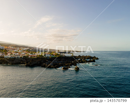 Coastal town strung along a black volcanic shoreline. Wide drone view of low white houses following a black volcanic shoreline on the western coast of Tenerife. The Atlantic opens out empty across the Coastal town strung along a black volcanic shoreline. Wide drone view of low white houses following a black volcanic shoreline on the western coast of Tenerife. The Atlantic opens out empty across the 143570799