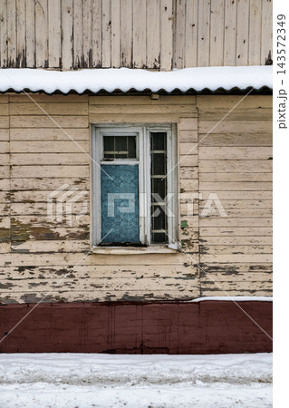 A wall of an old wooden house shows a blue window with cracked glass. Snow covers the ground beneath it. This scene is set in winter 143572349