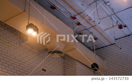 Ceiling lamps suspended beside exposed air conditioning ducts and a circular diffuser, fire sprinkler pipes and electrical conduits beneath concrete ceiling above white brick walls inside a building. Ceiling lamps suspended beside exposed air conditioning ducts and a circular diffuser, fire sprinkler pipes and electrical conduits beneath concrete ceiling above white brick walls inside a building. 143572730