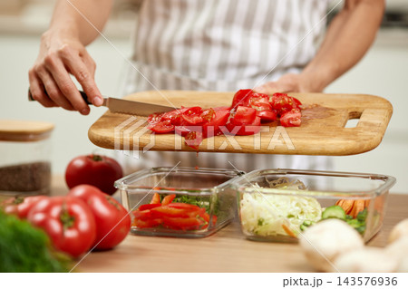 Person chopping fresh tomatoes on a cutting board, filling glass containers with vegetables for healthy meal preparation Person chopping fresh tomatoes on a cutting board, filling glass containers with vegetables for healthy meal preparation 143576936